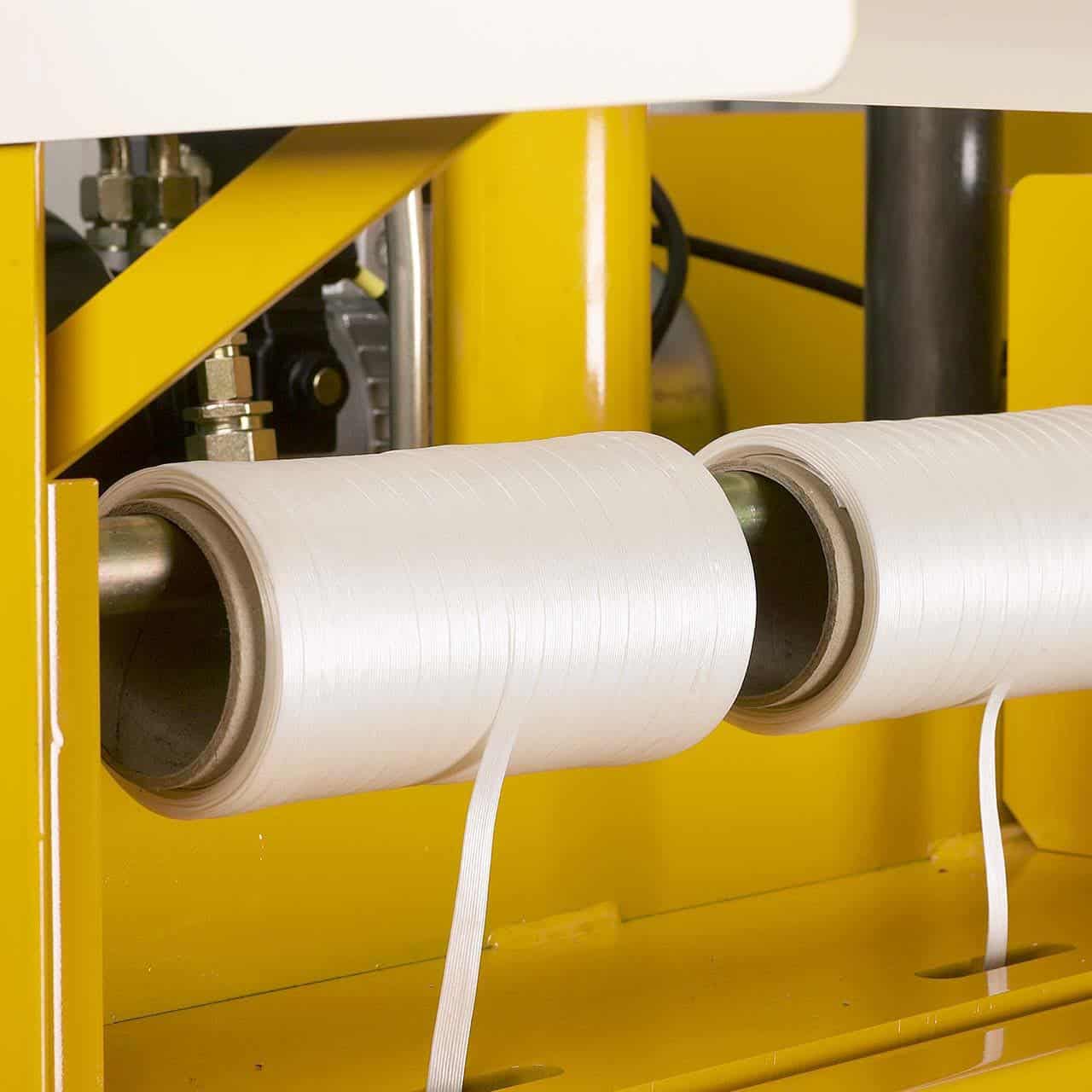 Close-up of a machine with two rolls of white baler strap being fed through yellow-colored components.